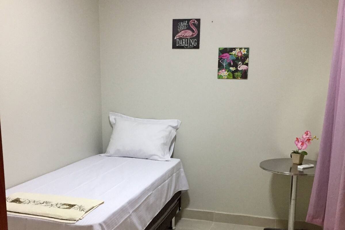 Ein kleines, ordentlich eingerichtetes Schlafzimmer mit einem Einzelbett, einem kleinen Tisch mit Blumen in einer Vase und einer Wand-Klimaanlage.