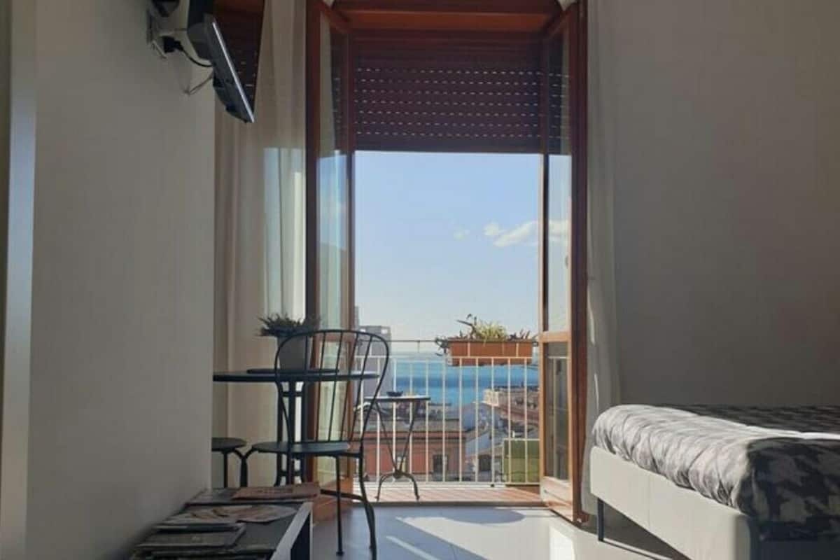 A small, modern room with a bed, a TV mounted on the wall, a table with chairs, and a balcony with a view of the sea.