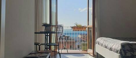 A small, modern room with a bed, a TV mounted on the wall, a table with chairs, and a balcony with a view of the sea.