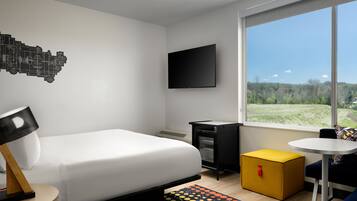 A modern hotel room with a bed, a TV, a small kitchenette, a window with a view of a green landscape, and a colorful rug.