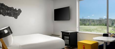 Una habitación de hotel moderna con cama, televisor, una pequeña cocina y un ventanal con vistas a un paisaje verde, además de una alfombra colorida.