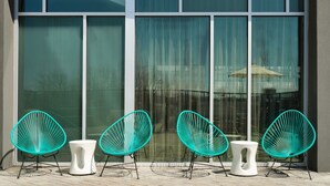 Un edificio moderno con ventanas amplias y un patio con cuatro sillas turquesa y dos taburetes blancos.