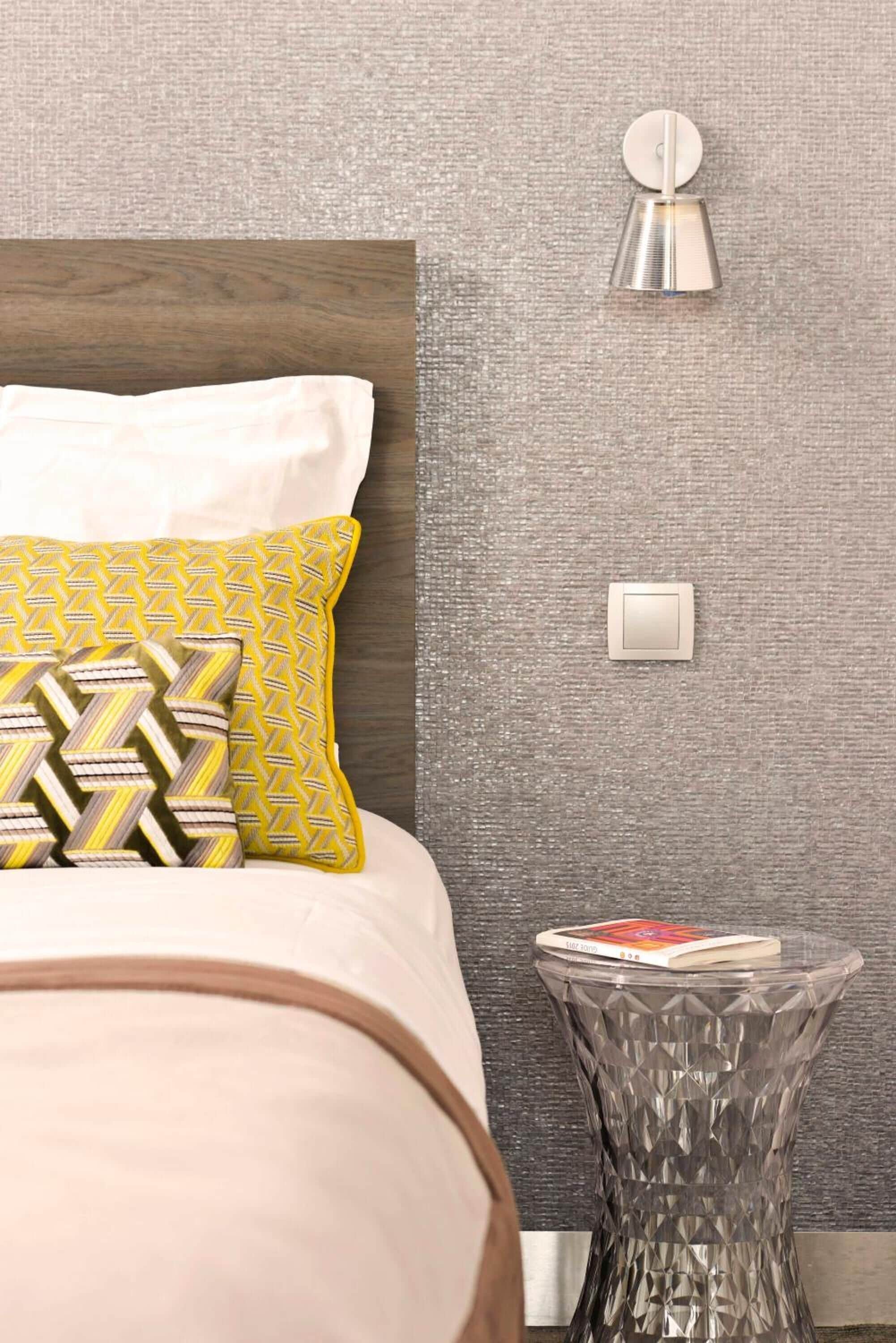 A modern bedroom with a bed, patterned pillows, a bedside table with a book, and a wall-mounted light fixture.