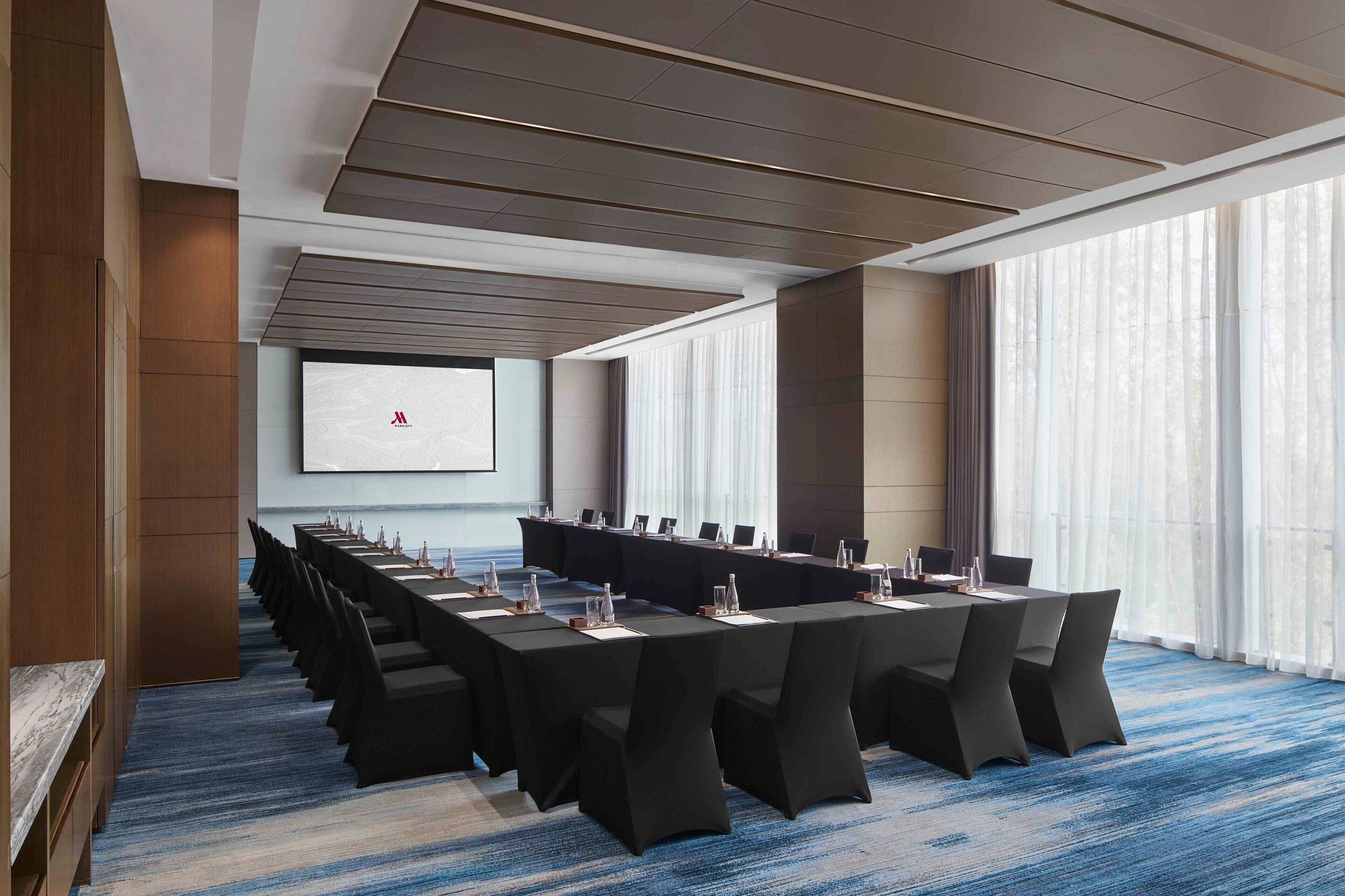 Un salón de conferencias con una larga mesa, sillas negras y una pantalla de proyector que muestra un logotipo.