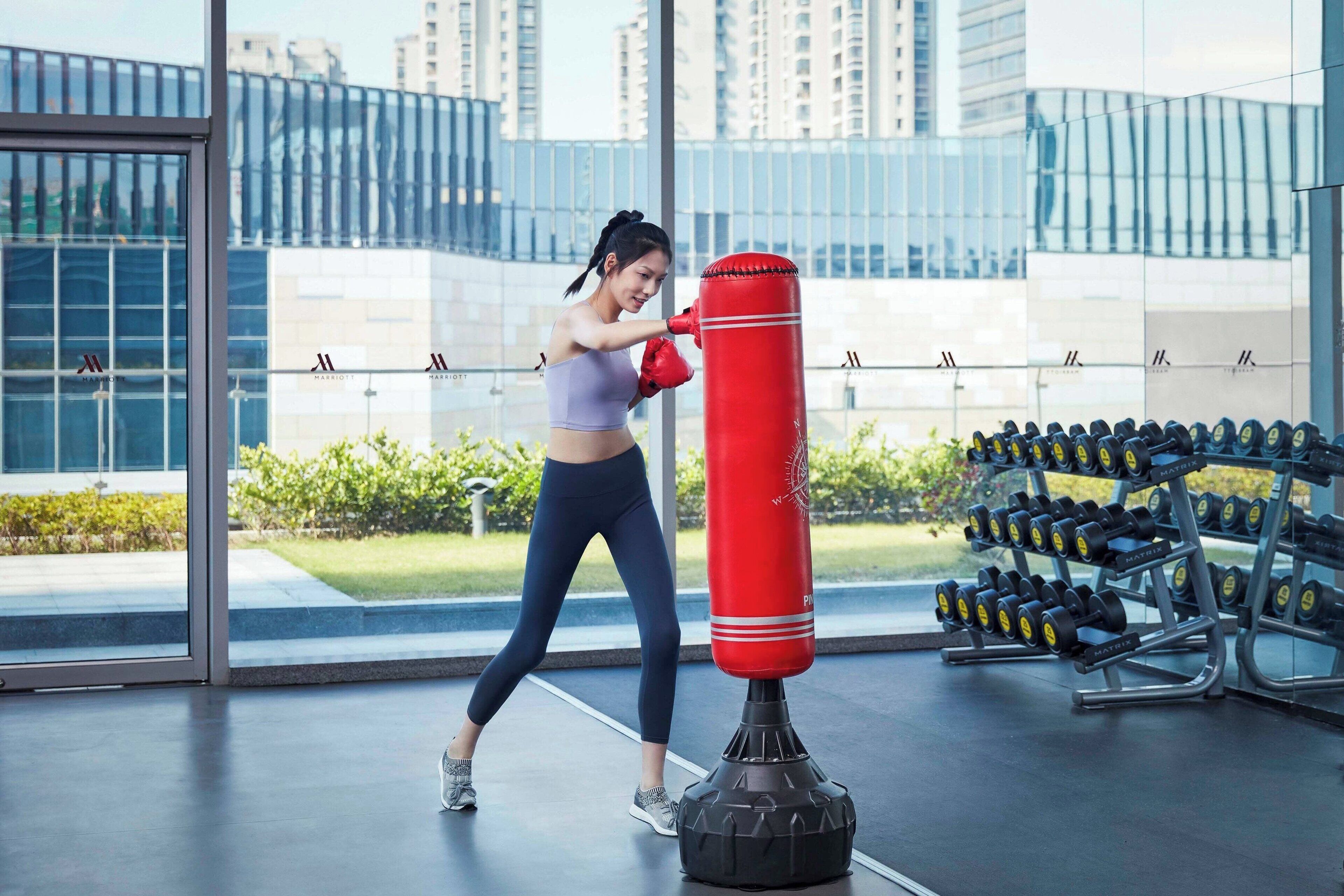 Une personne s’entraîne avec un sac de frappe dans une salle de sport moderne, dotée de grandes fenêtres et offrant une vue sur les bâtiments.