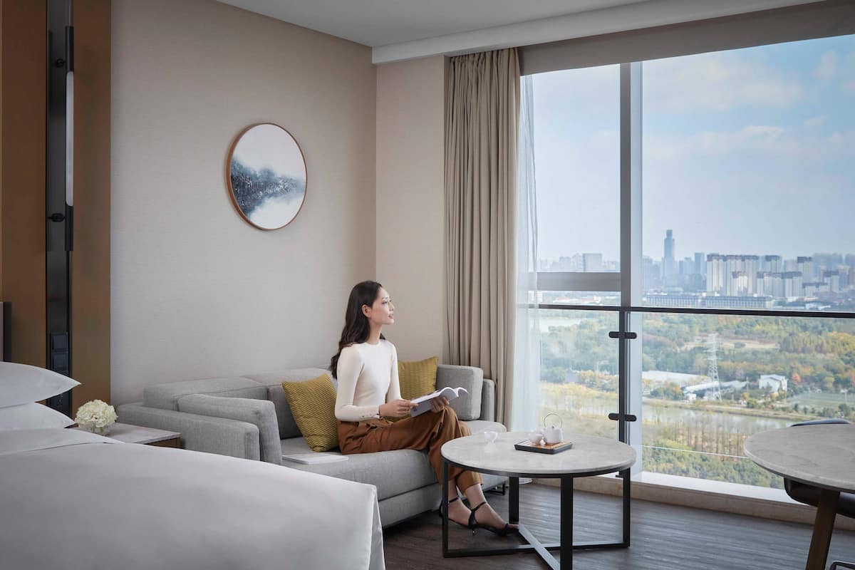 A modern hotel room with a large window offering a city view, a sofa, a coffee table, and a painting on the wall.