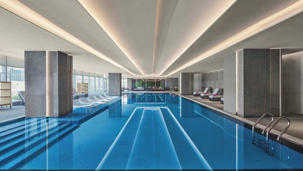 Una piscina interior moderna con carriles delimitados, sillas de descanso y vistas a la ciudad a través de amplios ventanales.