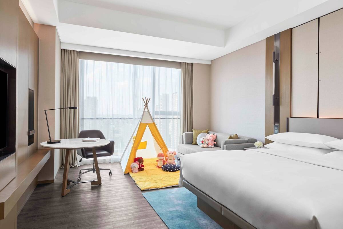 A hotel room with a bed, a desk, a chair, a small table, a window with curtains, a teepee, and a sofa with pillows.