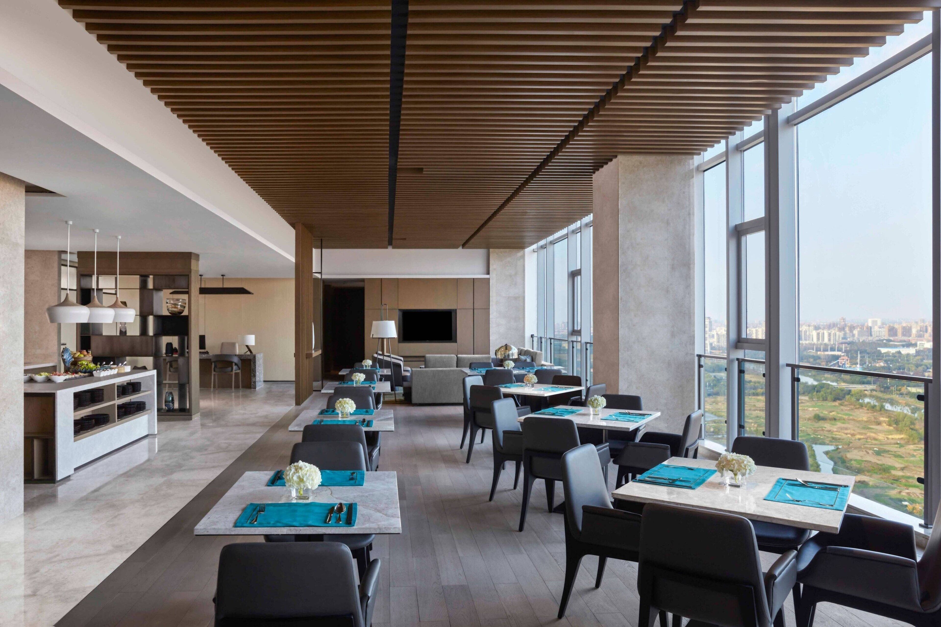 A modern dining area with tables set for a meal, large windows offering a city view, and a kitchen in the background.
