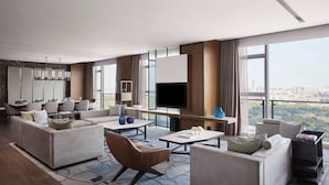 Un moderno salón de hotel con un amplio sofá, una mesa de centro y un televisor de pantalla plana. Hay una zona de comedor con una mesa y sillas, y vistas a la ciudad a través de amplios ventanales.