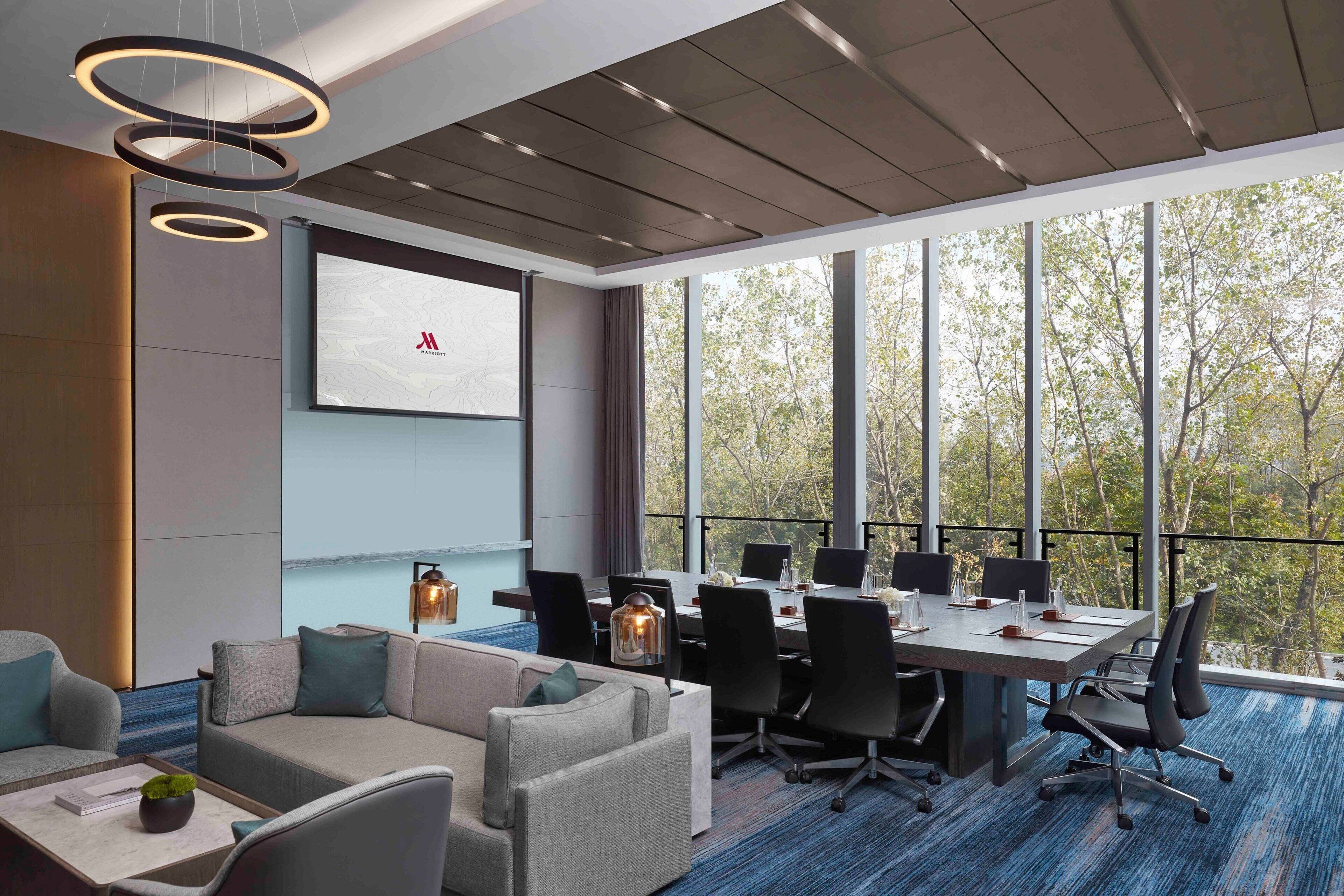 A modern conference room with a large table, comfortable seating, and large windows offering a view of trees.