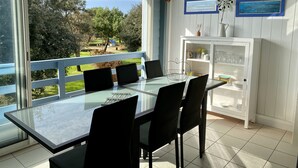 Ein Essbereich mit Glastisch und schwarzen Stühlen, ein weißes Schrank mit Glastüren sowie Blick auf Garten und Haus durch große Fenster.