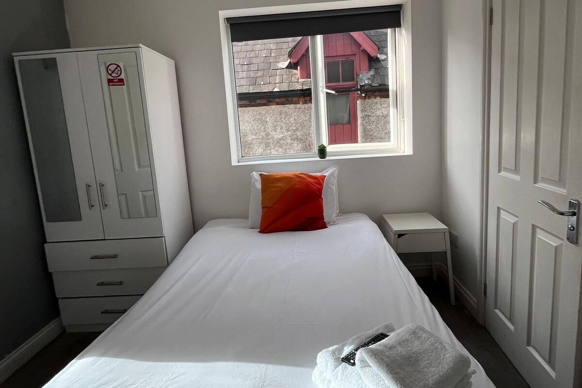 A neatly made bed with a white comforter and a red pillow, a white wardrobe, a small bedside table, and a window with a view of a building outside.
