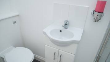 Ein Badezimmer mit einer weißen Toilette, einem weißen Waschbecken mit chromfarbenen Wasserhahn und einer rosa Flasche in einem Wandhalter.