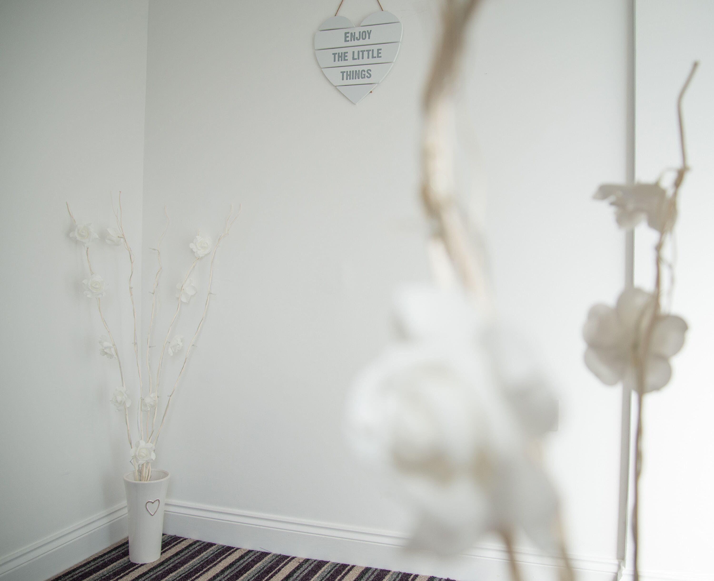 A white wall with a heart-shaped sign saying "ENJOY THE LITTLE THINGS" and a white vase with dried branches and flowers.