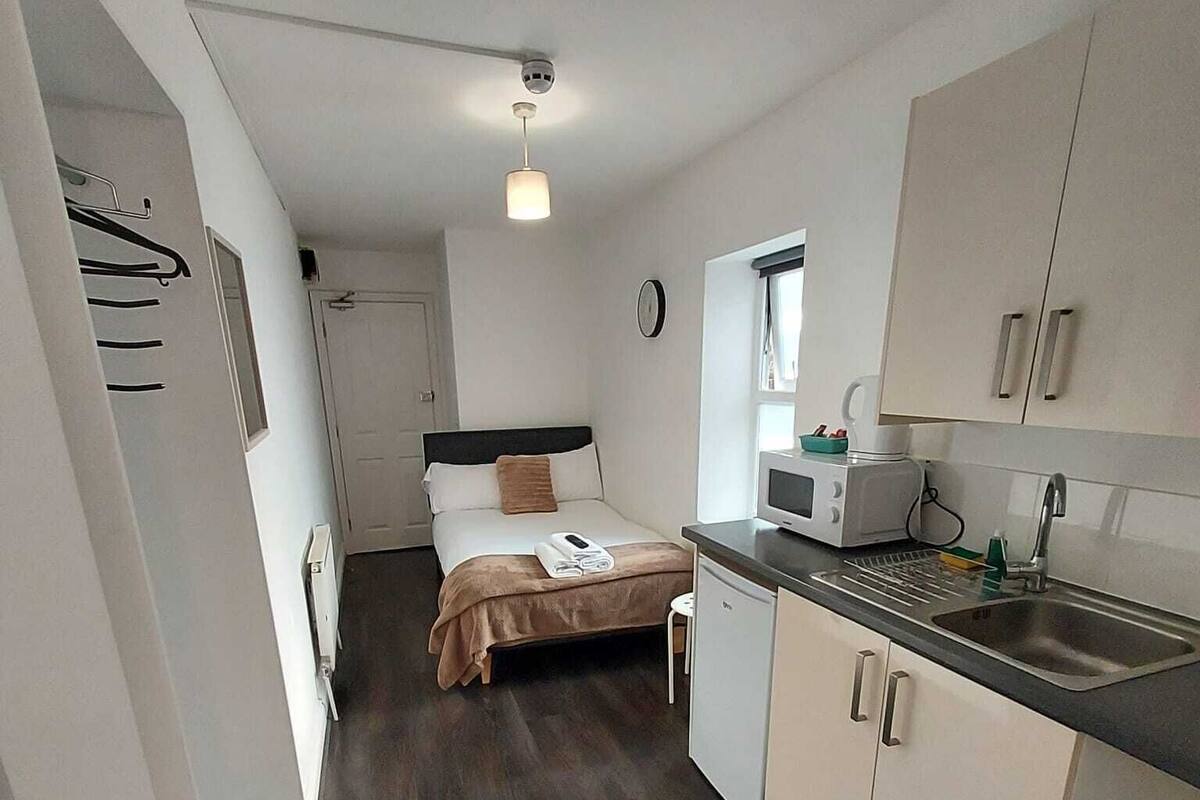 A compact hotel room with a bed, a small kitchenette, a ceiling fan, and a window.