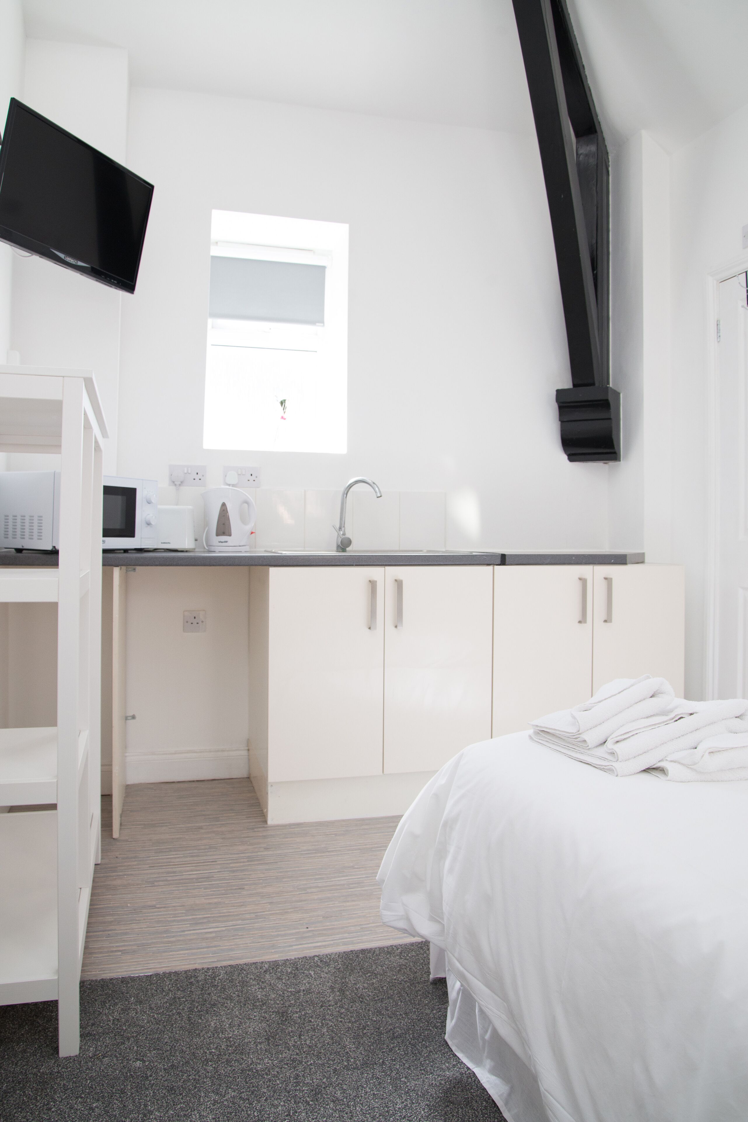 Un dormitorio moderno con cama, armario blanco, televisor integrado y zona de cocina con microondas, tostadora y lavabo.