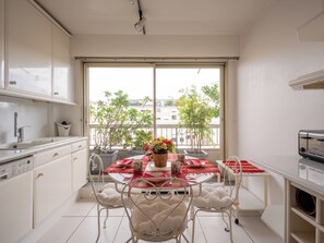 A modern kitchen with white cabinetry, a dining table with a red tablecloth, and a view of trees and buildings through a large window.
