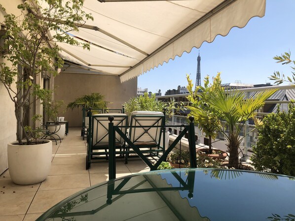 A rooftop terrace with potted plants, outdoor furniture, and a view of a tower in the background.