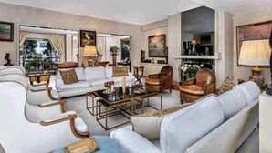 Un amplio salón con un gran sofá, una mesa de centro y una chimenea. Hay un ventanal con cortinas y un cuadro en la pared.