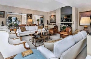 Ein großzügiges Wohnzimmer mit einer großen Couch-Garnitur, einem Couchtisch und einem Kamin. Es gibt ein großes Fenster mit Vorhängen und ein Bild an der Wand.