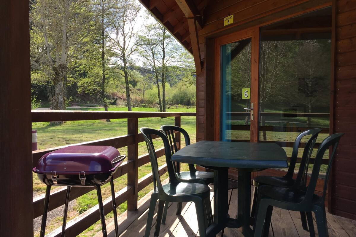 Une terrasse en bois avec une table et des chaises, un barbecue et une porte vitrée donnant sur une zone boisée.