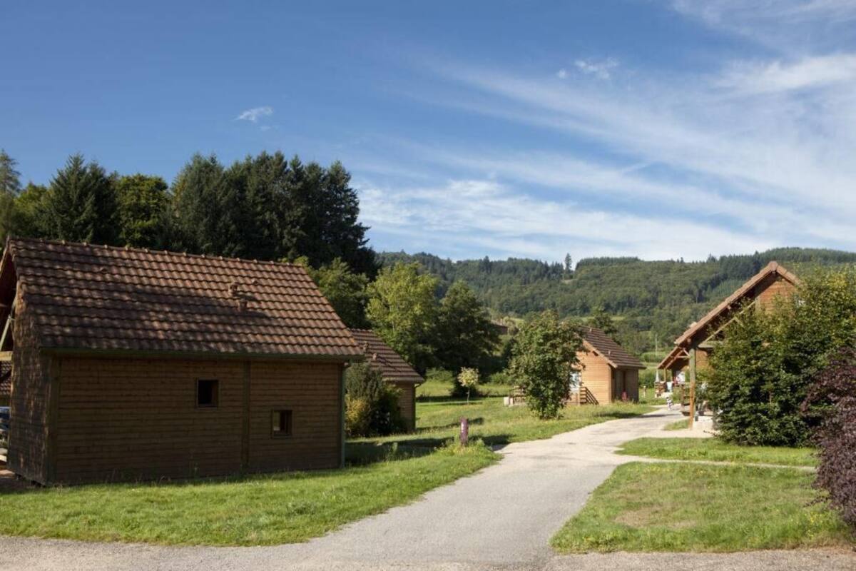 Un paysage rural avec des cabanes en bois, un chemin de gravier et une colline boisée en arrière-plan.