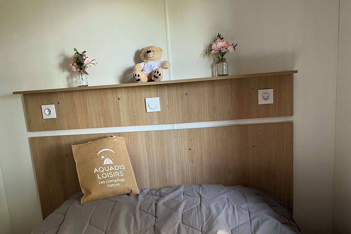 Un lit bien fait, avec une couverture grise, une tête de lit en bois, une petite étagère ornée d’un ours en peluche et de fleurs, et un oreiller avec un motif imprimé.
