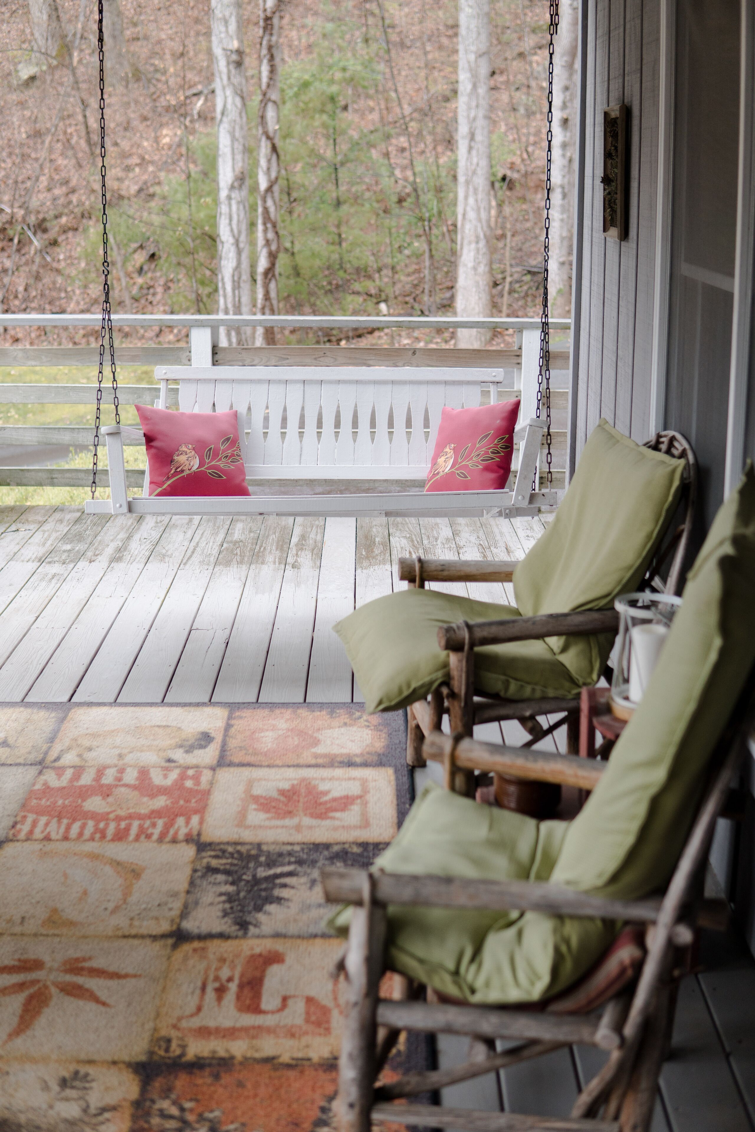 A wooden porch with a swing and two rocking chairs, a white fence, and a patterned rug.
