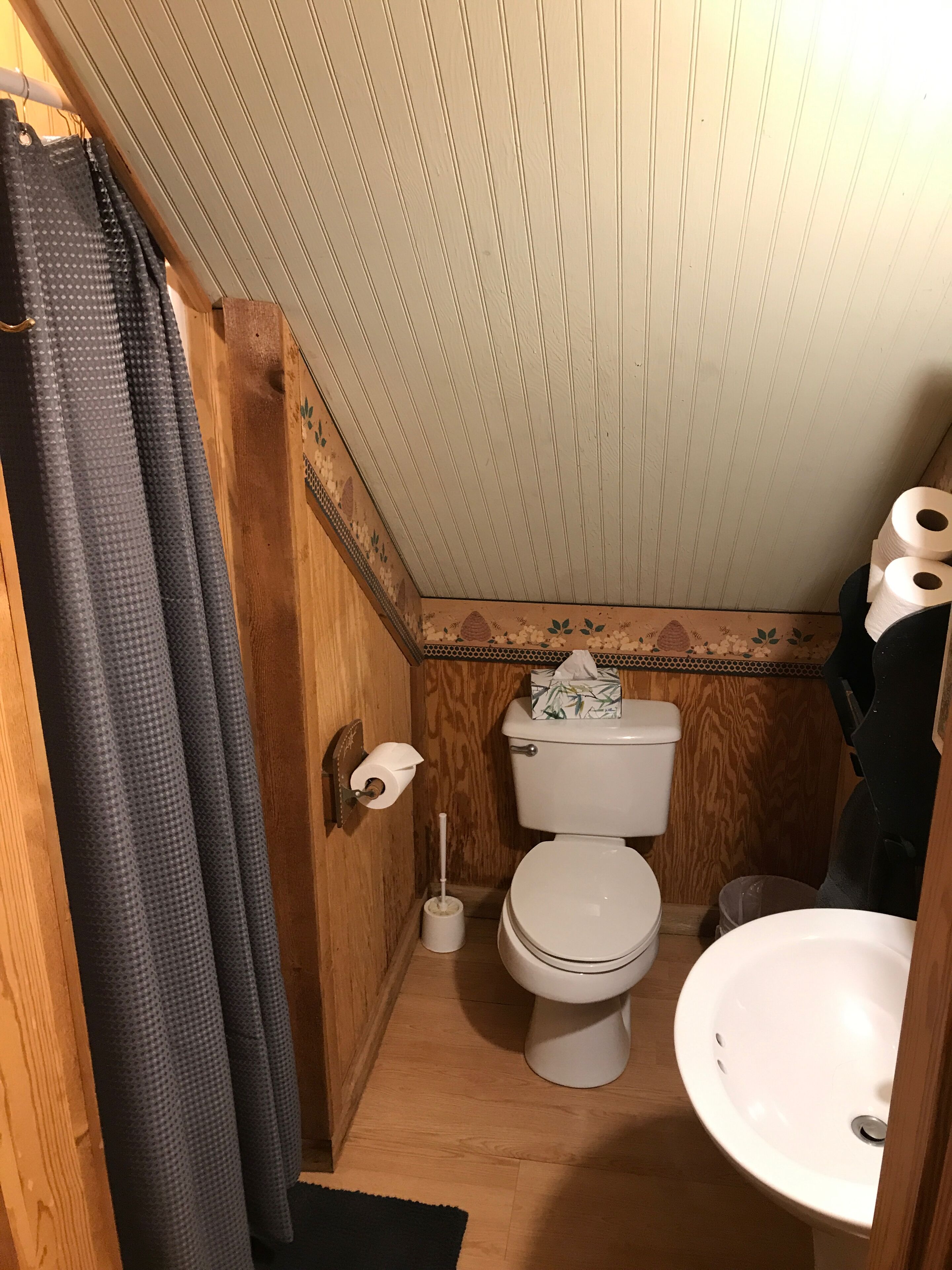 Une petite salle de bain aux dimensions réduites, avec un plancher en bois, des toilettes blanches, un lavabo blanc et un rideau de douche.