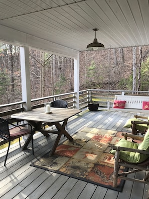 Een overdekt terras buiten, met een tafel en stoelen, omringd door een houten balustrade en met uitzicht op een bebost gebied.