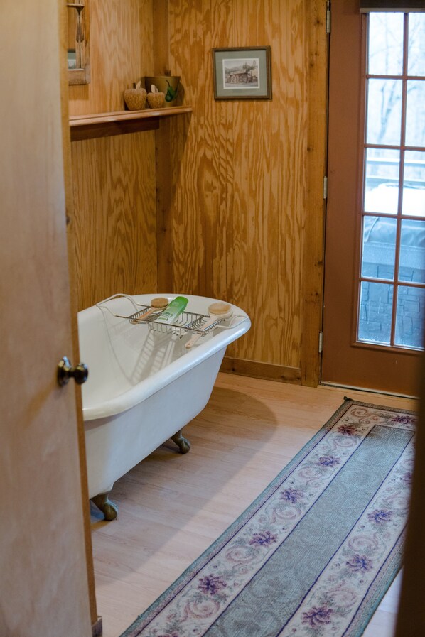 A bathroom with a wooden wall, a white bathtub, a small shelf with toiletries, and a window with a view of trees.