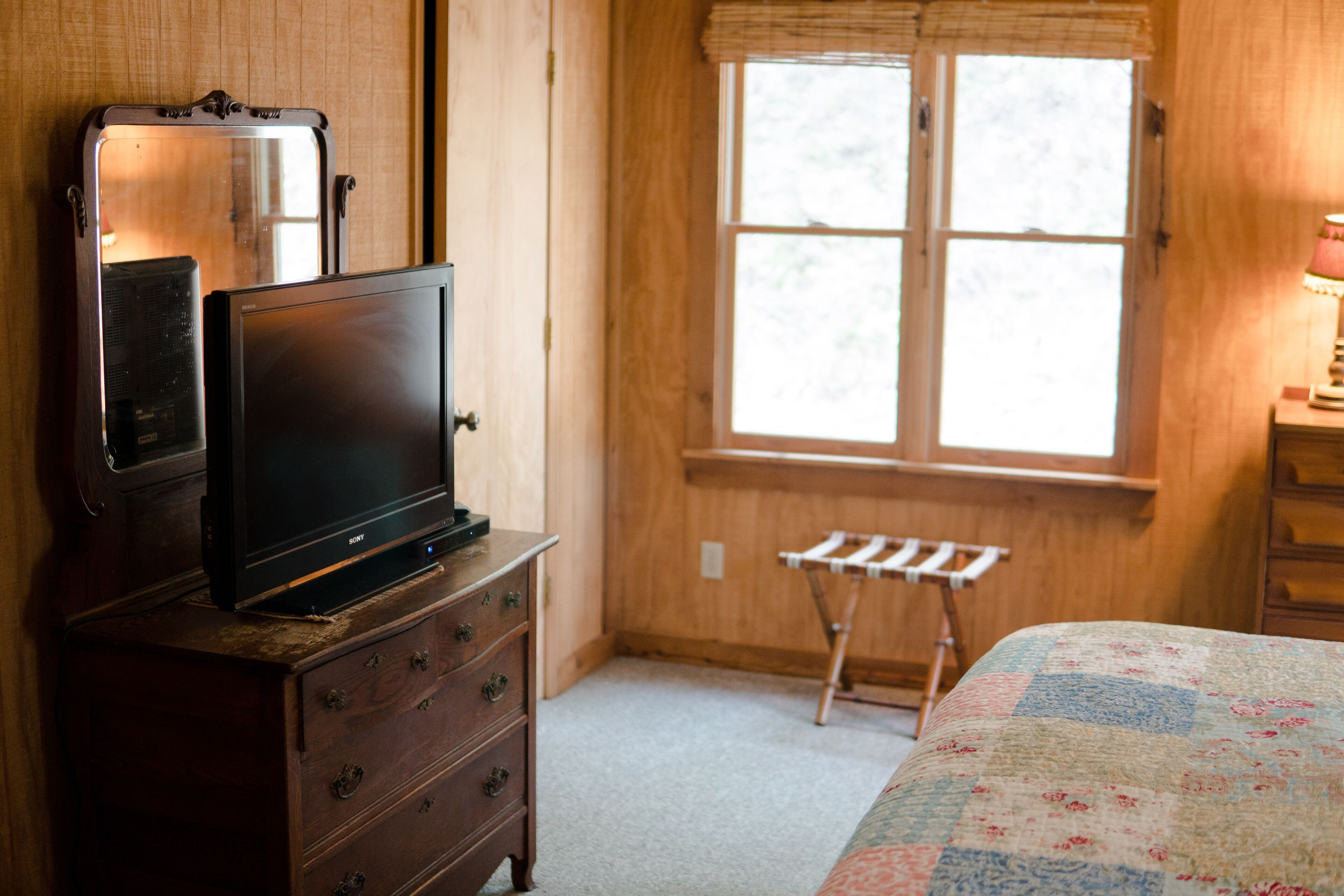 A bedroom with a wooden dresser, a television on top, a window with a view of trees, and a bed with a quilt.