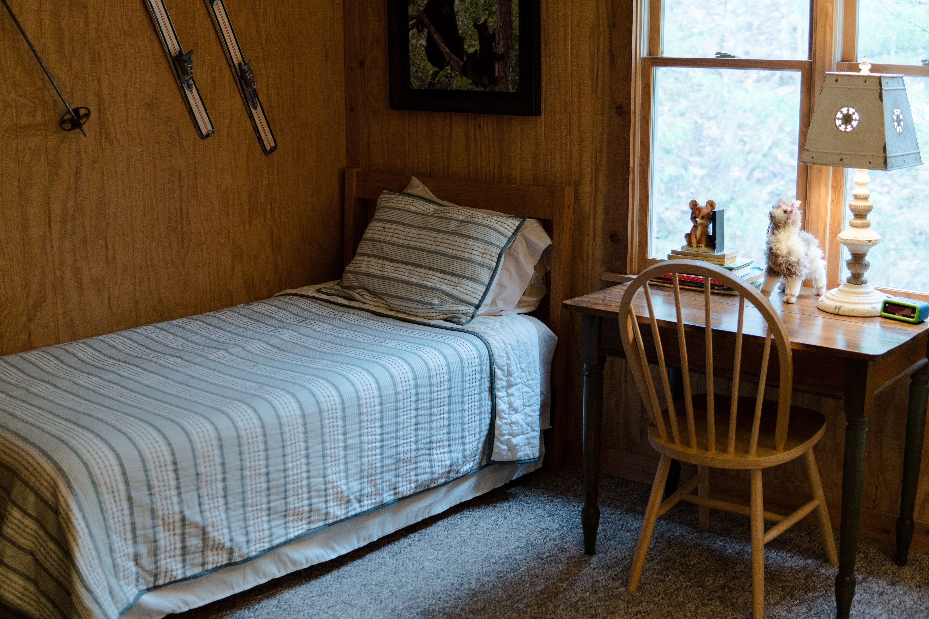 A small, cozy room with a single bed, a wooden desk, and a chair. There are skis hanging on the wall and a lamp on the desk.