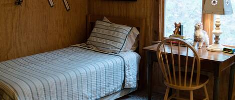 A small, cozy room with a single bed, a wooden desk, and a chair. There are skis hanging on the wall and a lamp on the desk.