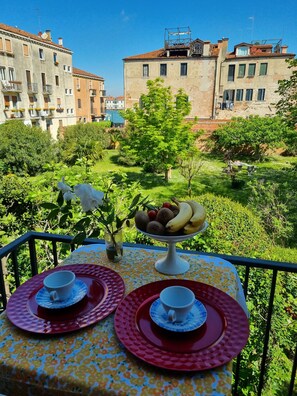 A balcony with a table set for two, a fruit bowl, and a vase with a plant. The background features a garden and buildings. - RELAX GARDEN HOUSE WITH LAGOON VIEW (VENEZIA)