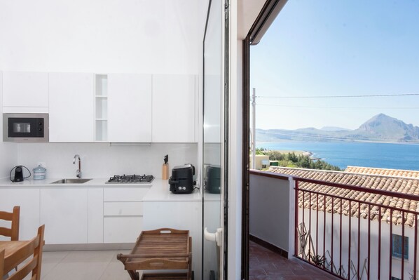 A compact kitchen with a sink, stove, and cabinets, a wooden table, and a balcony with a view of the sea and mountains.