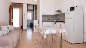 Uma cozinha moderna com armários brancos, uma geladeira branca e um micro-ondas. Há uma mesa de jantar com toalha de mesa estampada e cadeiras transparentes. Um sofá bege com almofadas estampadas está na sala de estar.