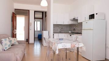 Une cuisine moderne avec des meubles blancs, un réfrigérateur blanc et un four à micro-ondes. On y trouve une table à manger recouverte d’une nappe à motifs et entourée de chaises transparentes. Dans l’espace salon, il y a un canapé beige agrémenté de coussins à motifs.