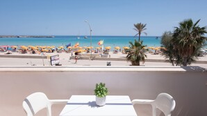 Varanda com mesa e cadeiras, vista para uma praia com guarda-sóis e pessoas.