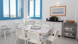 Comedor con una mesa preparada para cuatro personas, sillas blancas, una encimera de cocina con electrodomésticos y vistas a través de las ventanas.