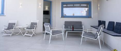 Uma área de estar moderna ao ar livre, com cadeiras brancas e cinzentas, uma mesa pequena e um banco.