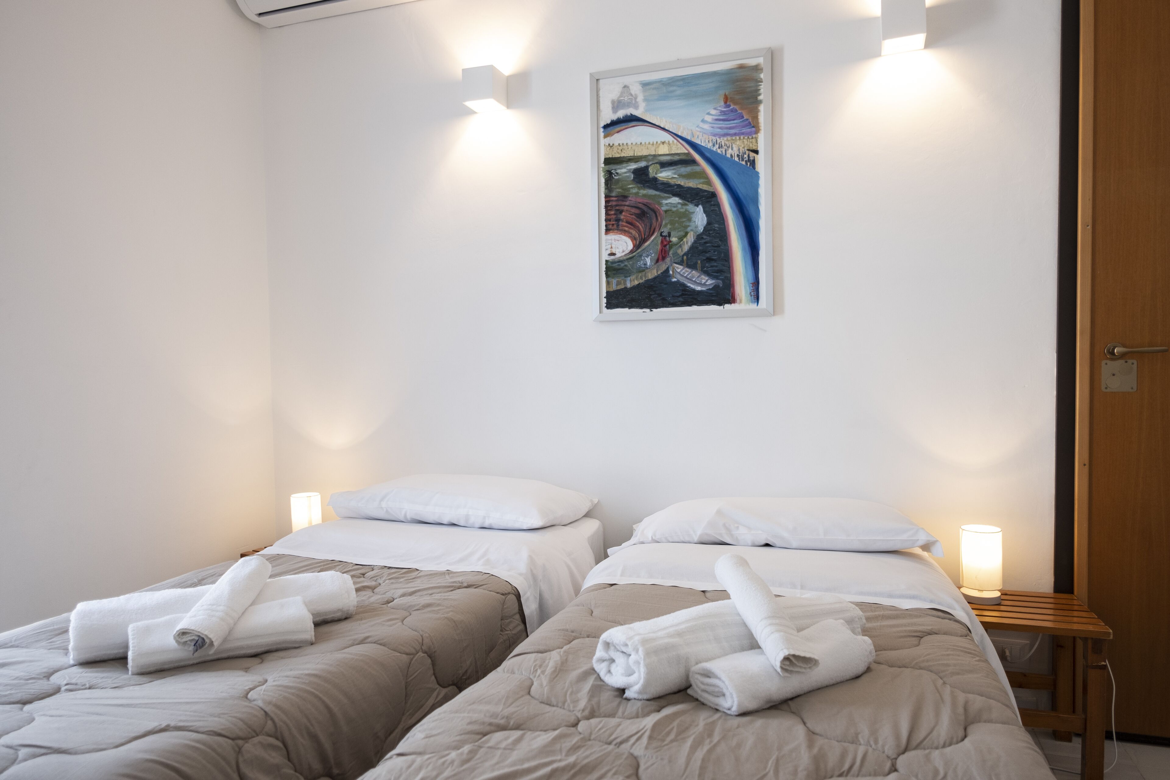 A hotel room with two beds, each with a beige quilt and white pillows, and a painting on the wall.