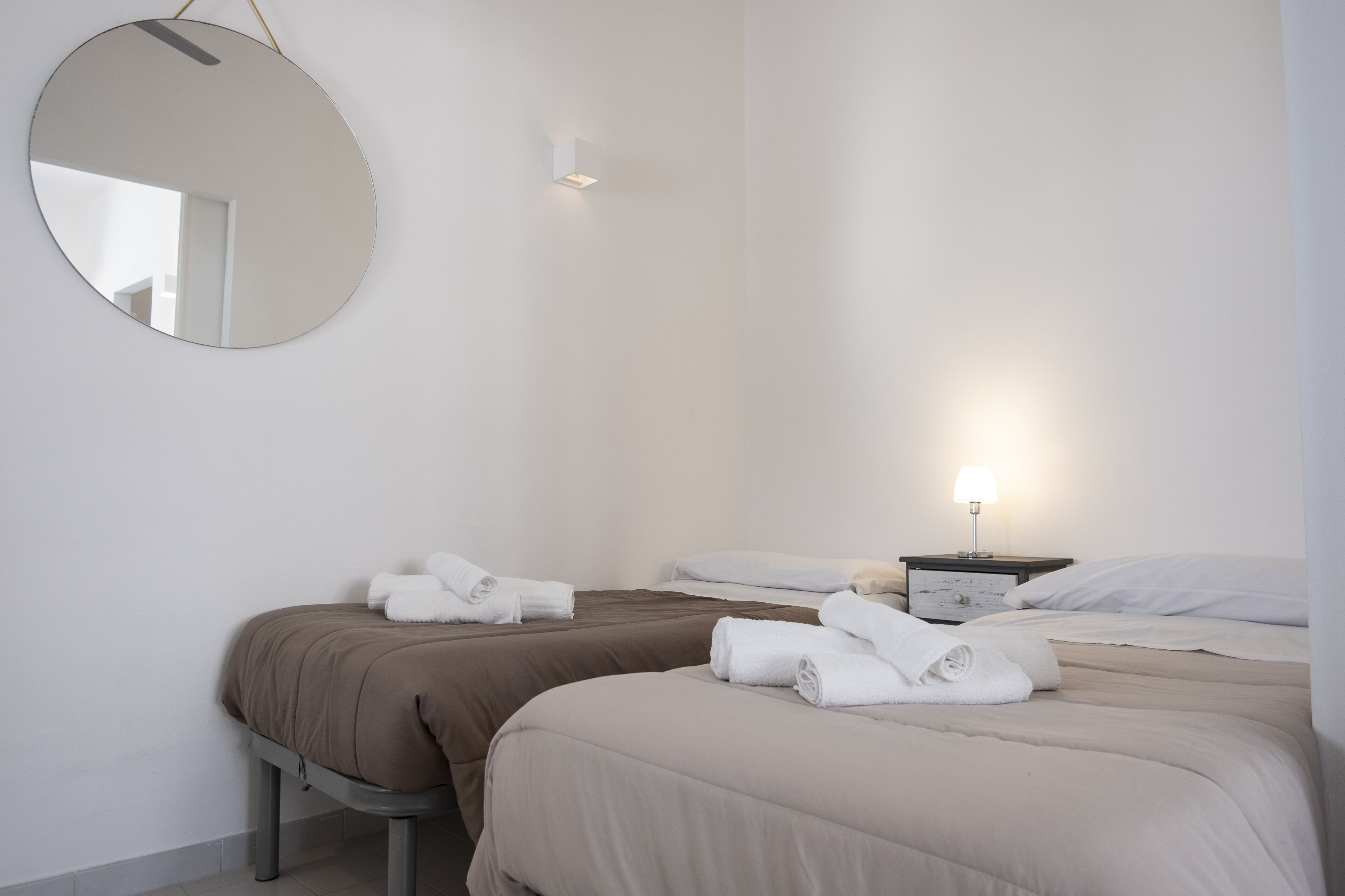 A hotel room with two beds, a round mirror, a bedside table with a lamp, and neatly folded towels.