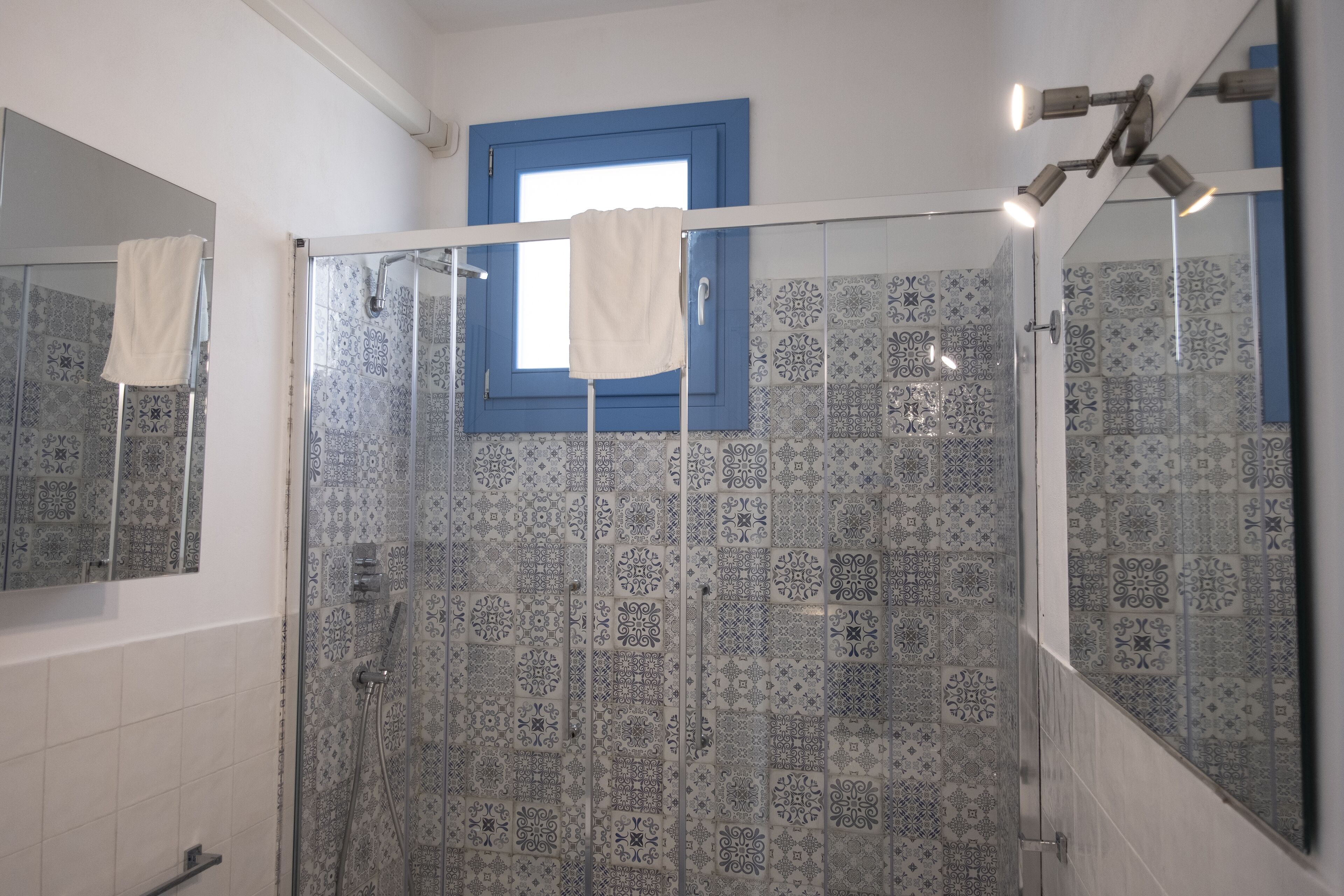 A bathroom with patterned tiles, a glass shower enclosure, a white towel hanging on the shower door, and a blue-framed window.