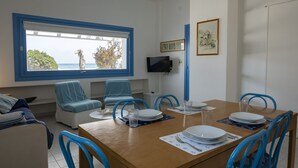 A dining area with a wooden table set for four, blue chairs, and a large window offering a view of the sea.