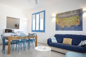A room with a wooden dining table, blue chairs, a blue sofa, a white round side table, a map on the wall, and a window with blue shutters.