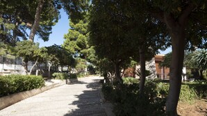 A paved pathway lined with trees and shrubs, leading to a building with a brick facade. - Villa Faro (San Vito Lo Capo)