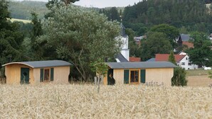 Two wooden cabins in a wheat field with a church and houses in the background. - Schäferwagen am Berghof (Zeulenroda-Triebes)