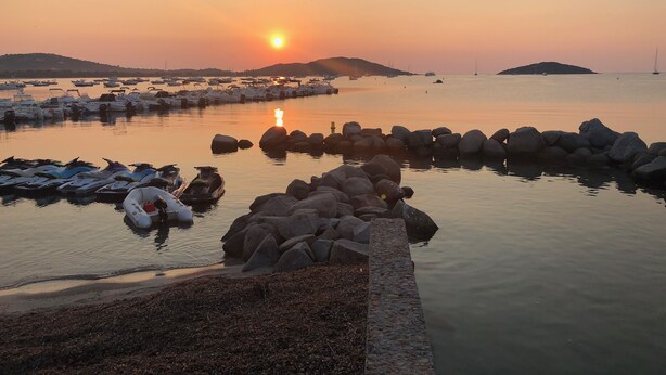 Una marina con barche ormeggiate, un molo roccioso e un tramonto sullo sfondo.